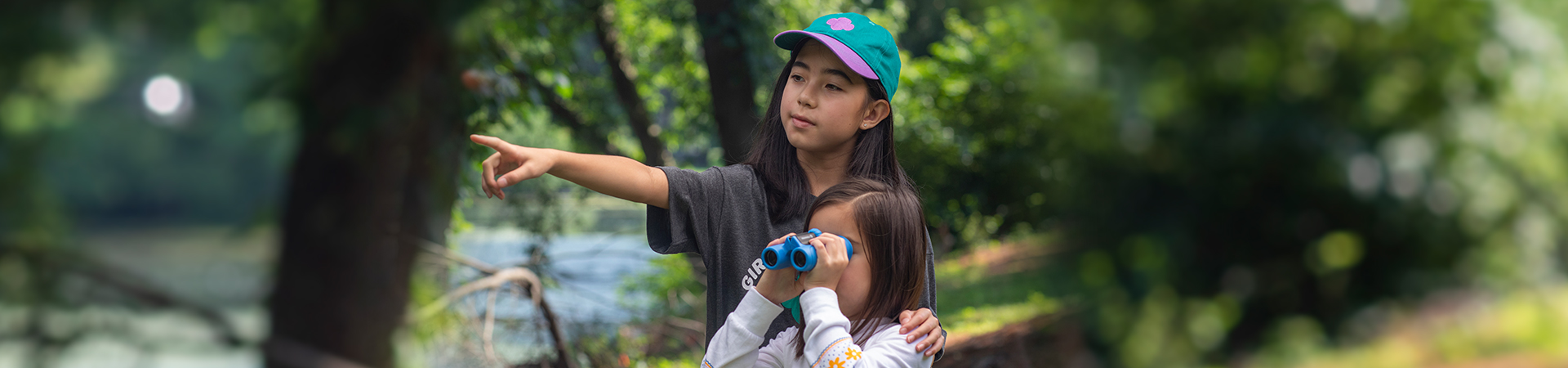  A Girl Scout points ahead and a younger Girl Scout looks through binoculars. 